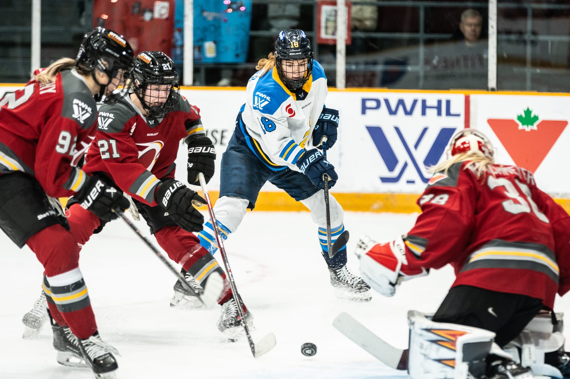 Two Ottawa players (both left) are going for the puck against the Toronto player (middle). Maschmeyer is on her knees in the goal (right). The Ottawa playersare in red, while the Toronto player is in white.