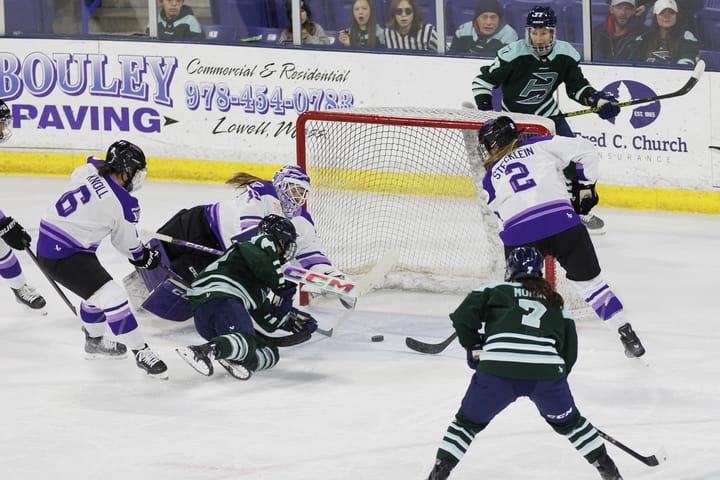 Saulnier lays on the ice right after pushing the puck towards the goal, and it's about to go past a diving Rooney. There are three other players from both Minnesota and Boston around the net. The Boston players are in green, while the Minnesota players are in white.