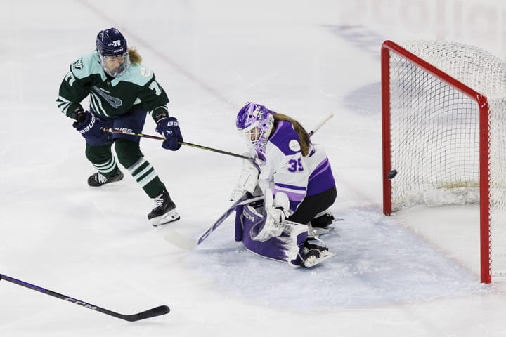 With her back to the goal, Tapani (left in green) points her stick toward the net to follow through on her shot. Rooney (right in white) is on her knees attempting to make the save, but the puck has sailed past her.