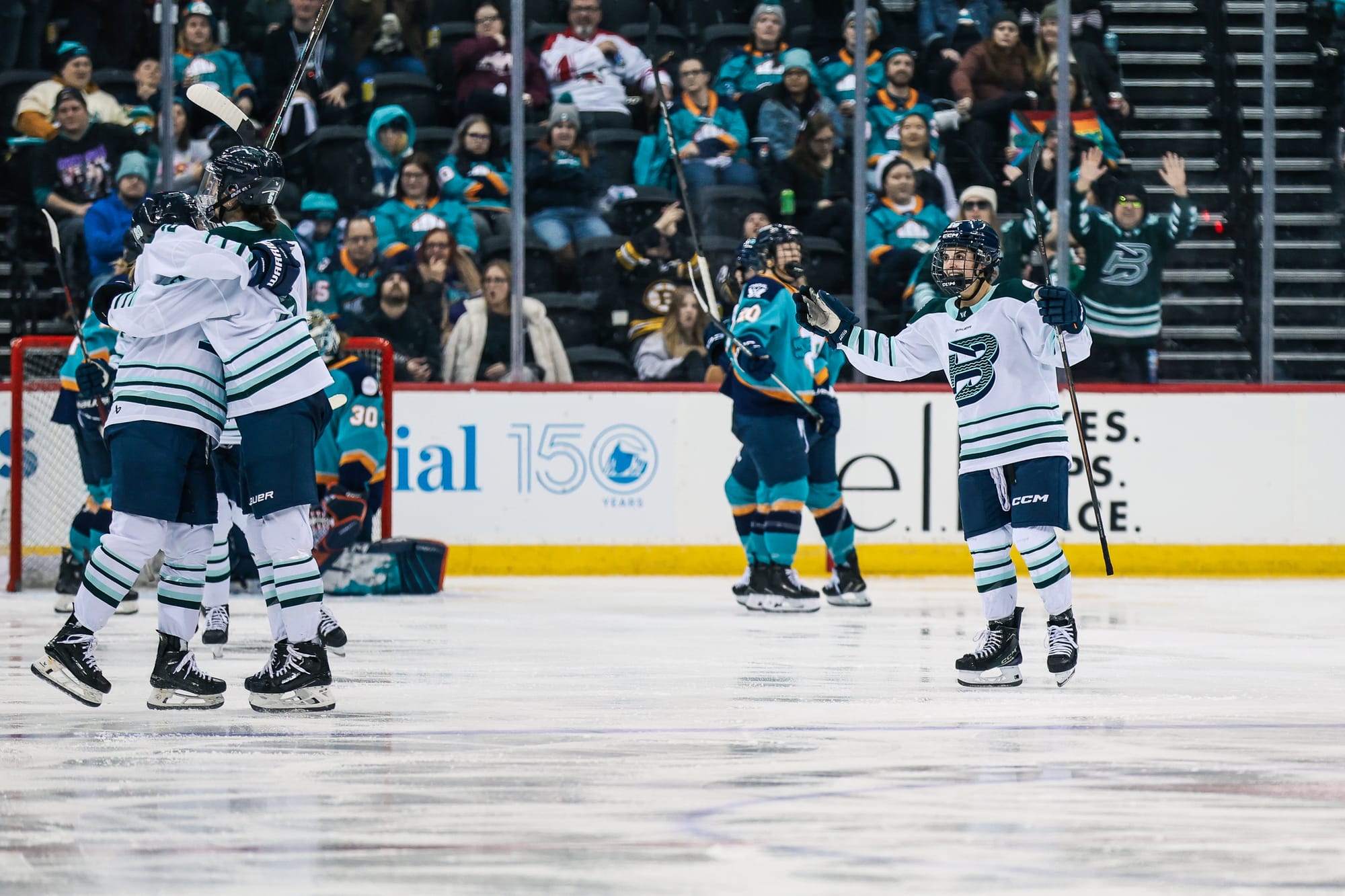 Two Fleet players celebrate with a hug on the left side of the frame. Another is skating in from the right to join. They are wearing white away uniforms.