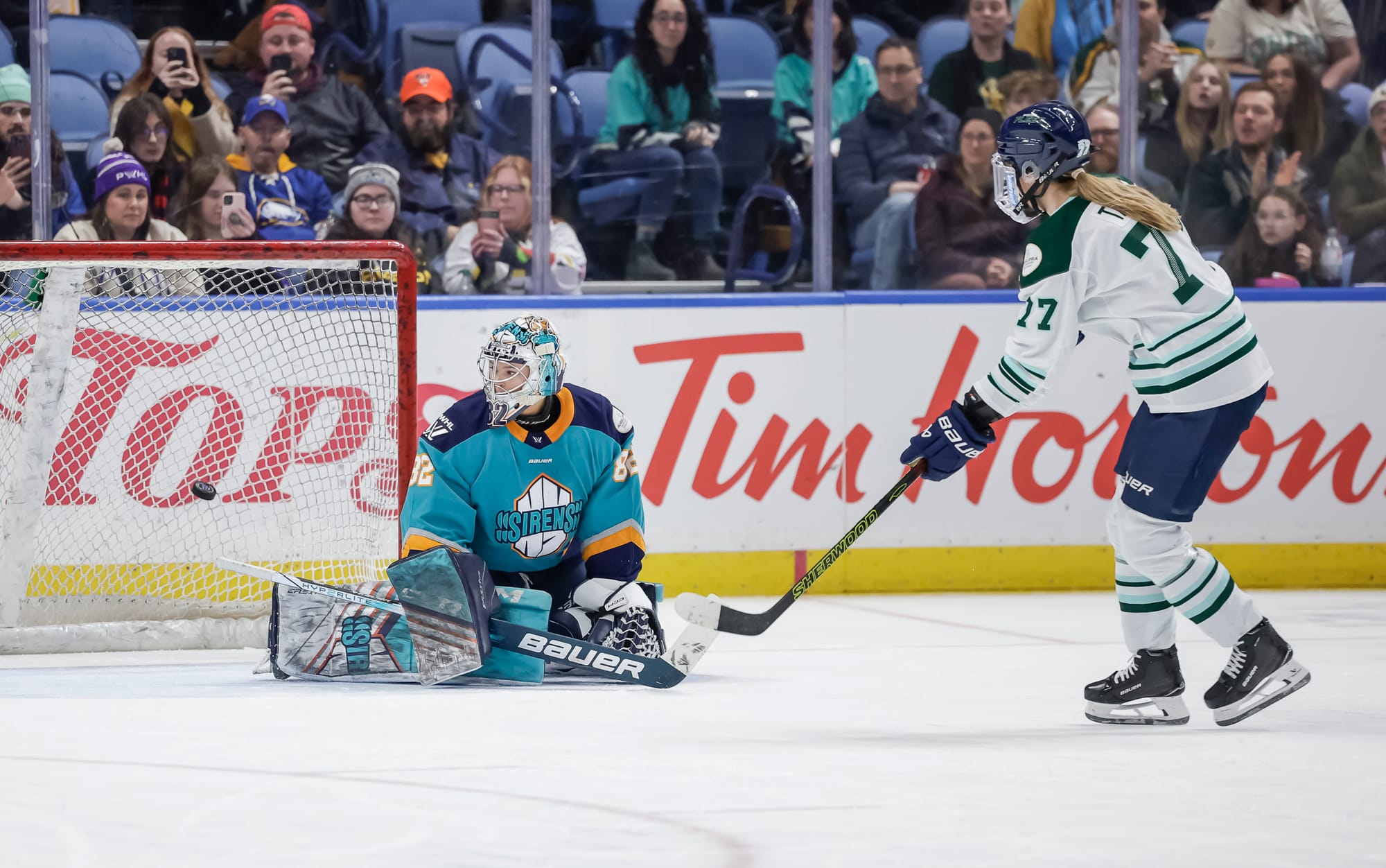 Tapani (right in white) has her stick slightly lifted and pointed towards the net after releasing her shot. Osborne (left in teal) is on her knees attempting to make the save, but the puck has sailed past her.