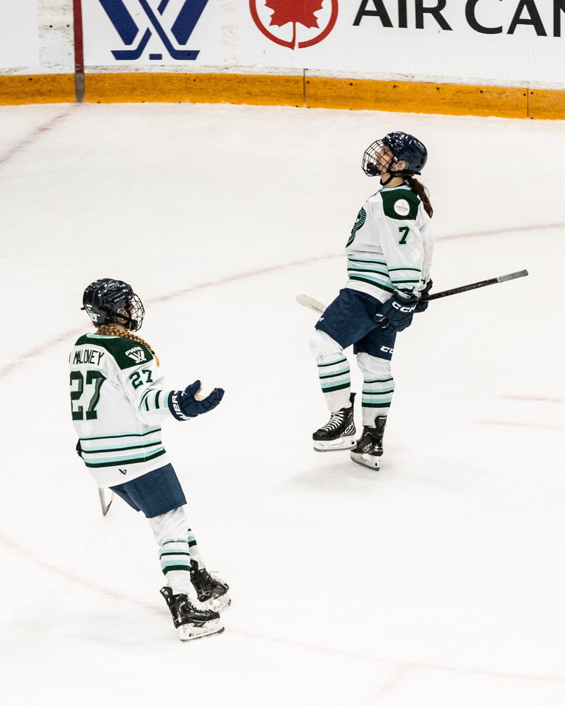 Morin pumps her fists in celebration, while Maloney skates in with her arms out for a hug. They are wearing white away uniforms.