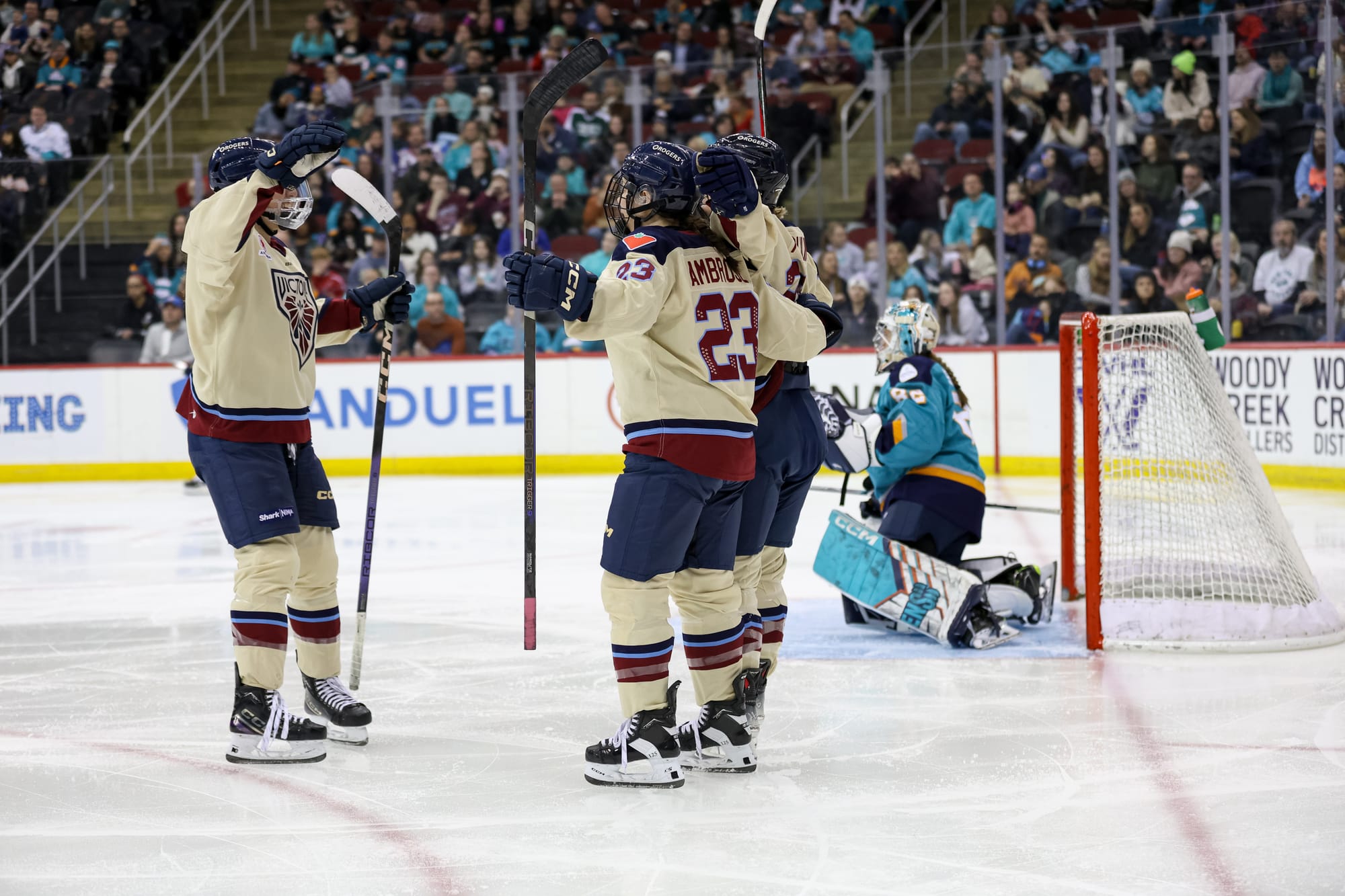 Three Victoire players celebrate a goal with a group hug. They are off to the side of the crease, and Sirens netminder Kayle Osborne is out of focus in the background. The Victoire are wearing cream away uniforms, while Osborne is in teal.