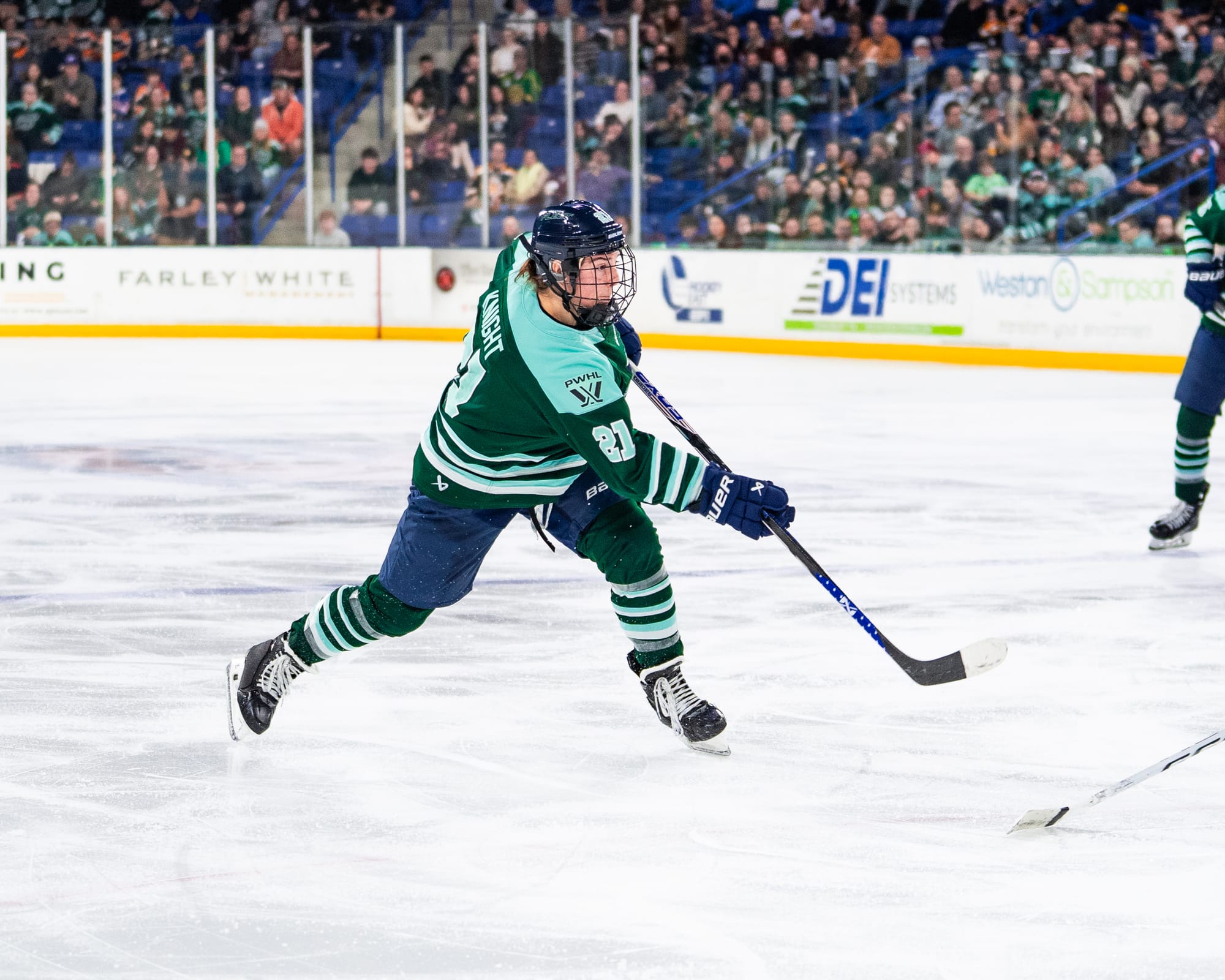 Knight looks ahead and shifts her weight to her left leg as she follows through on a shot. The puck has already left the frame. She is wearing a green home uniform. 