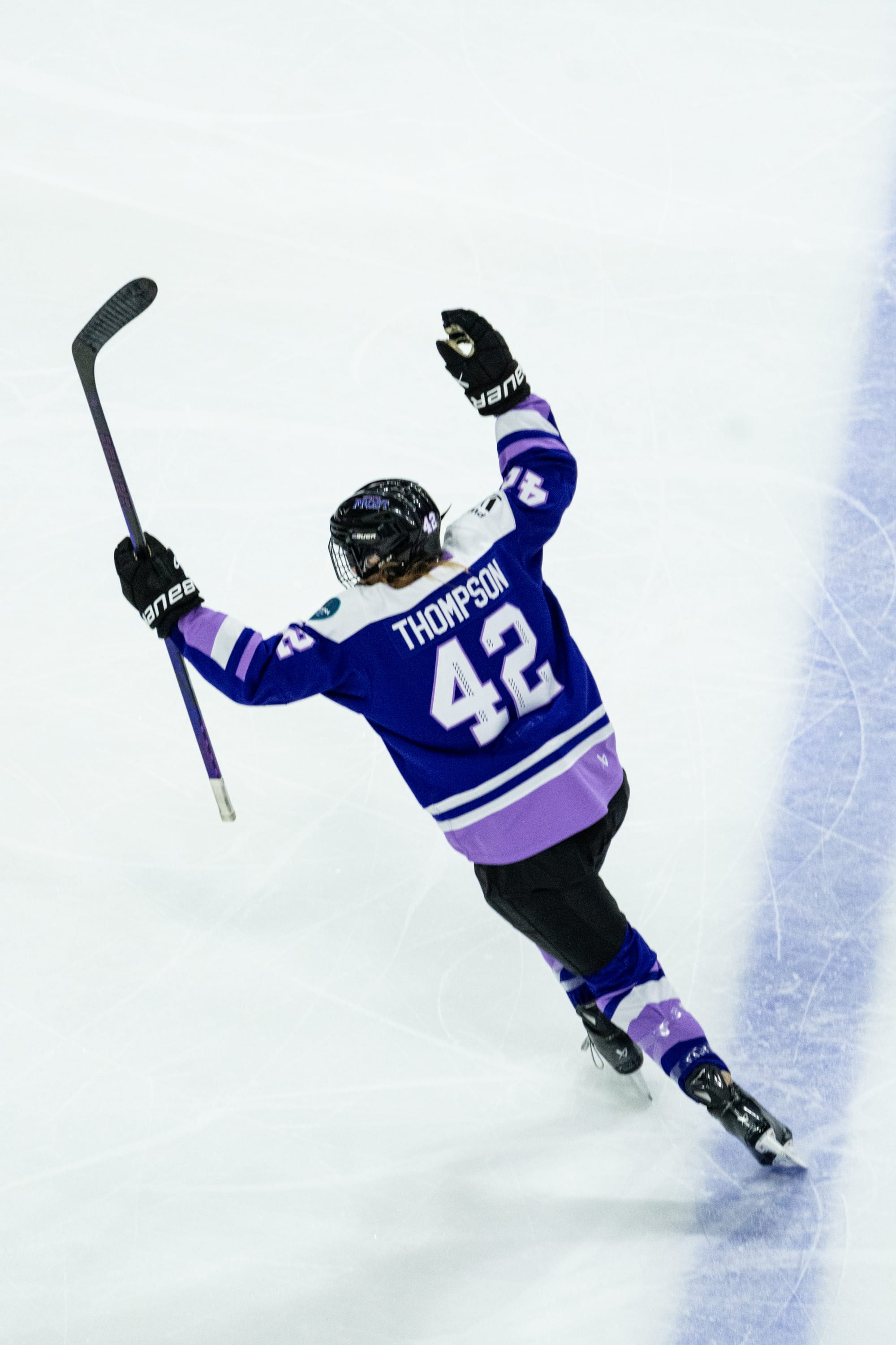 Thompson is back to the camera as she raises her arms in celebration as she skates away from the blueline. She is wearing a purple home uniform.