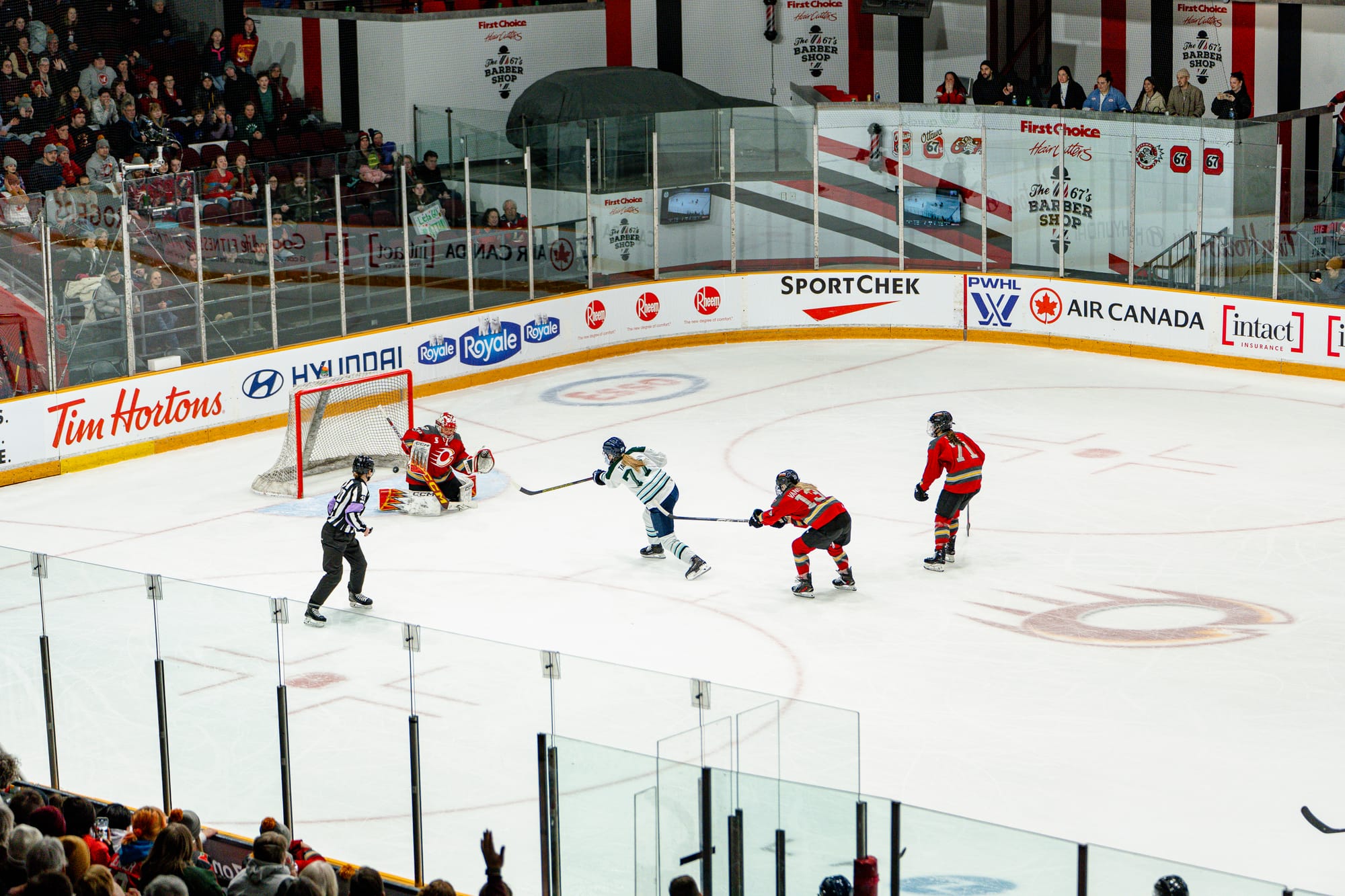 An above-ice view of Tapani, wearing white, following through on her shot. The puck is just passing Maschmeyer. Two Charge players, wearing red, are chasing Tapani. 