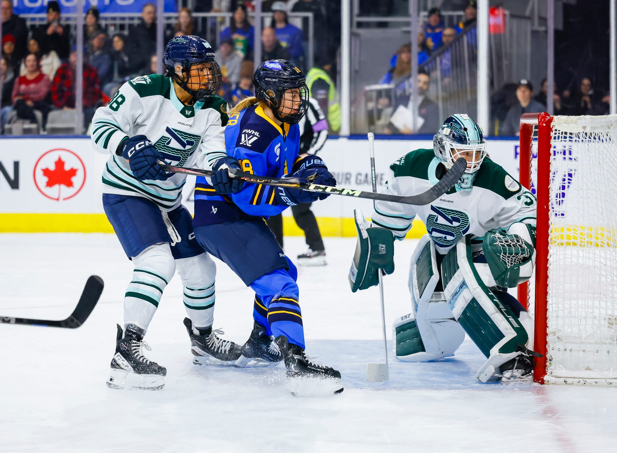 Adzija (left in white) and Compher (right in blue) battle right outside Aerin Frankel's crease. They are leaning into each other as they watch the puck, which is out of frame behind the goal.