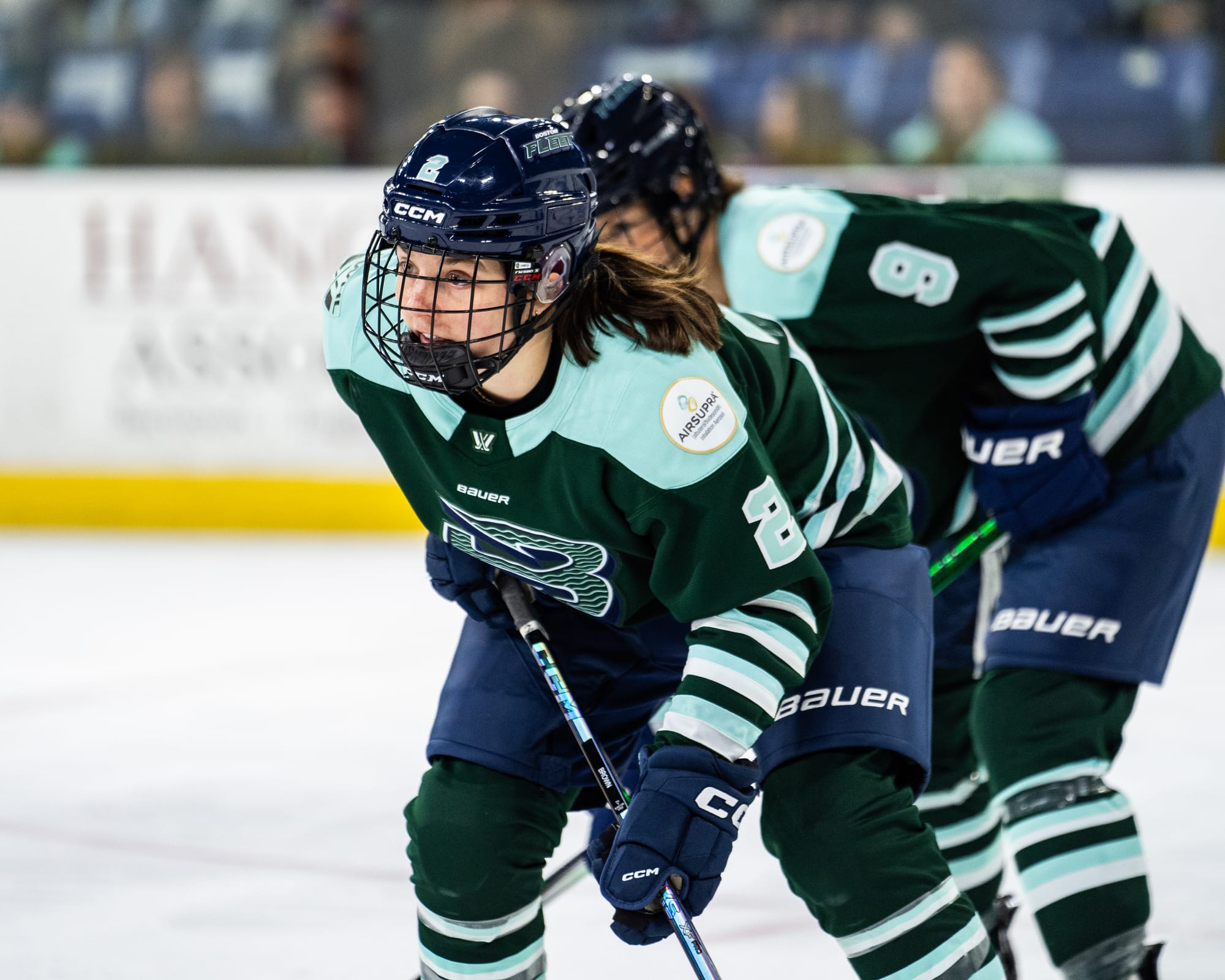 Brown looks up and crouches in a hockey stance at the hash marks of a circle. Sophie Shirley is behind her out-of-focus. They are wearing green home uniforms. 