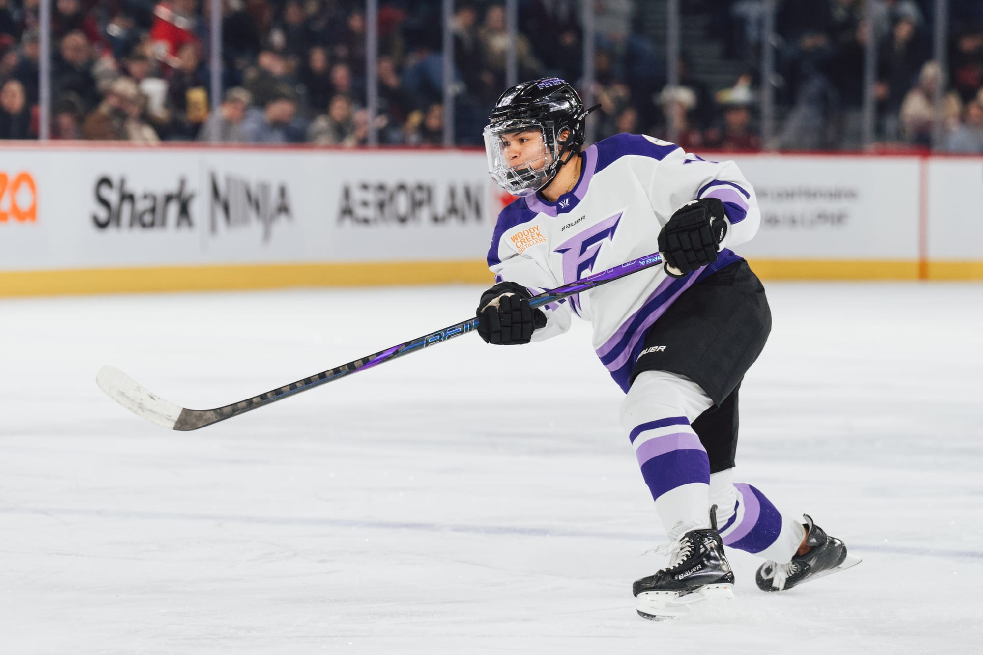 Jaques looks toward the net as she raises her stick to follow through on a shot. Her weight is primarily on her left leg as she stands near the blueline. She is wearing a white away uniform.