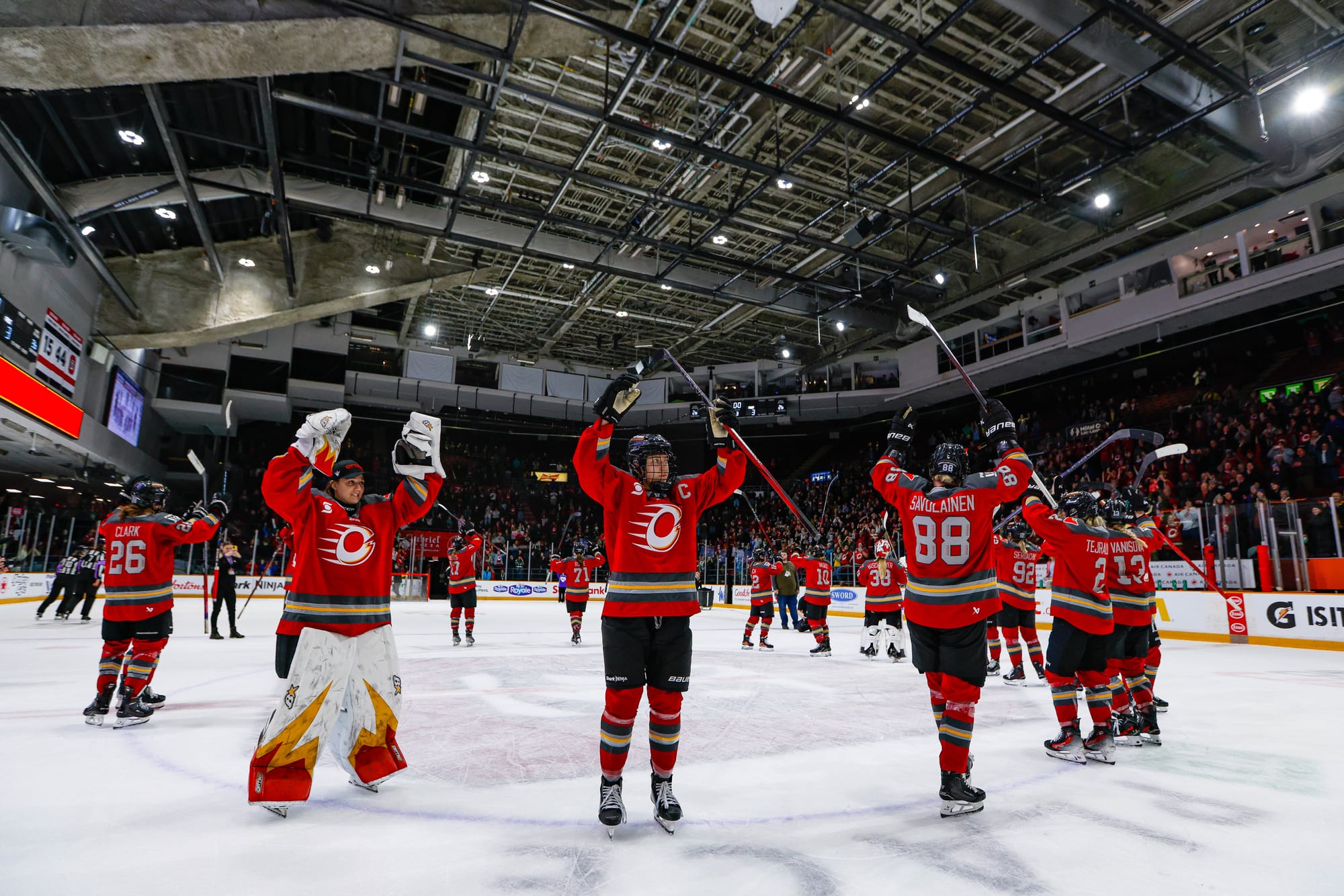 Charge players raise their hands above their heads while standing around the center ice circle. It is part of their postwin celebration. They are wearing red home uniforms.