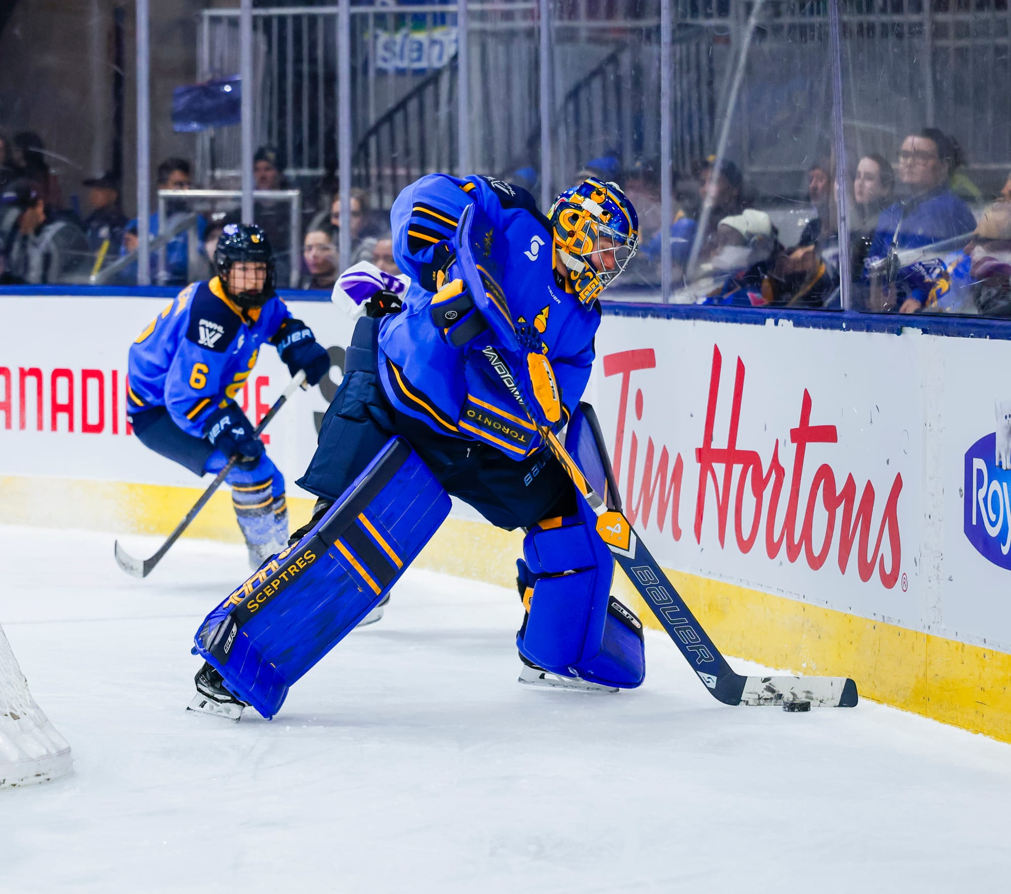 Kirk is directly behind her net, with the puck on her stick just off the end boards. She is looking down at is as she prepares to make a play, and wearing a blue home uniform.