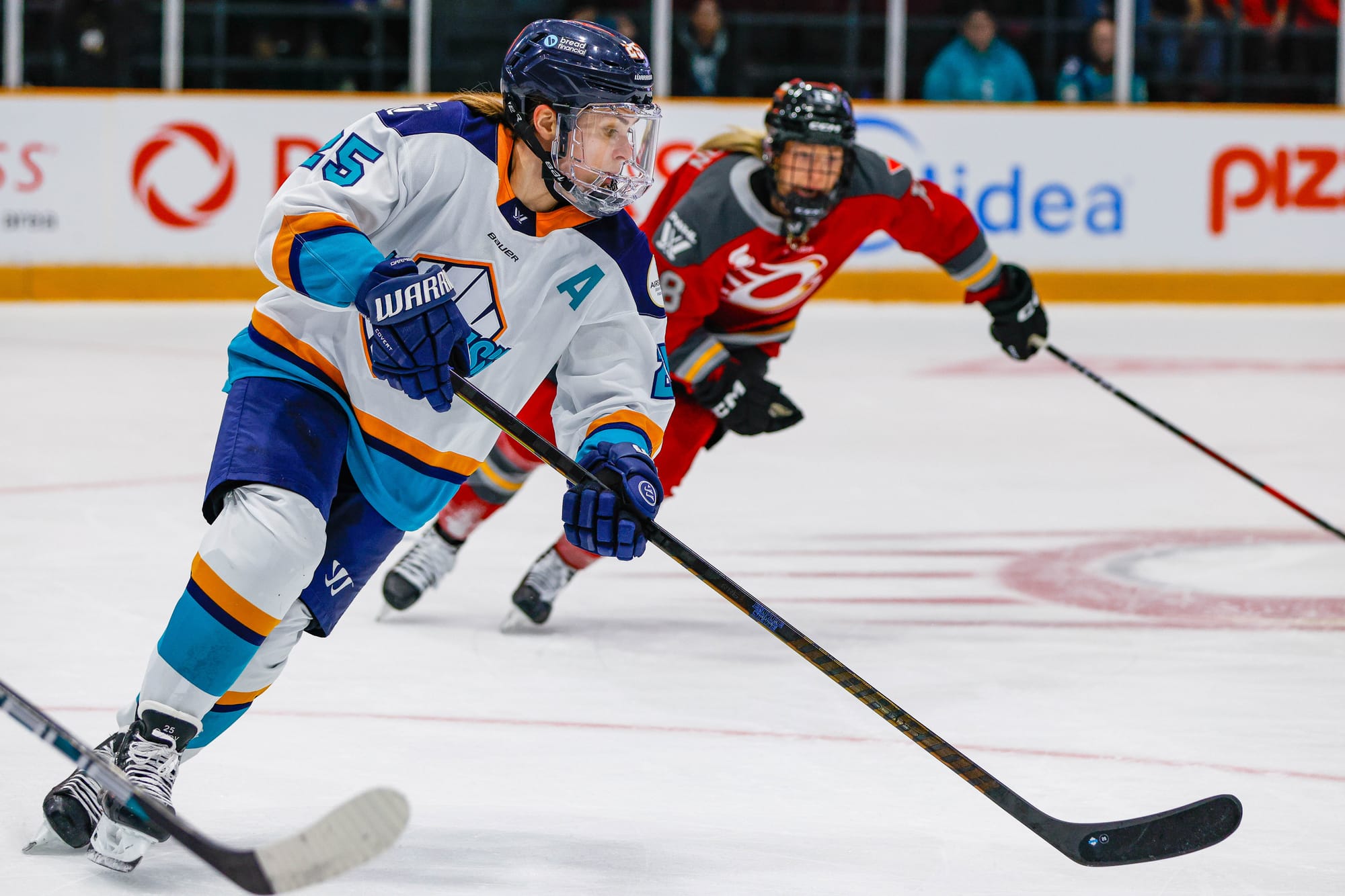 Carpenter looks down at the puck as she skates through one of the faceoff circles. She is wearing a white away uniform.