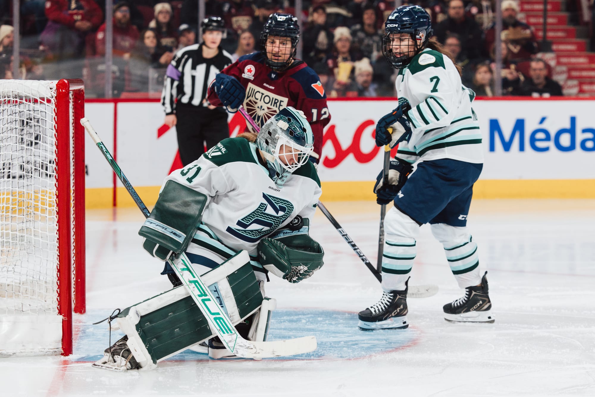 Frankel kneels in her crease and looks down at the puck in her glove. She is wearing a white away uniform. There is a Montréal and Boston skater in the background.