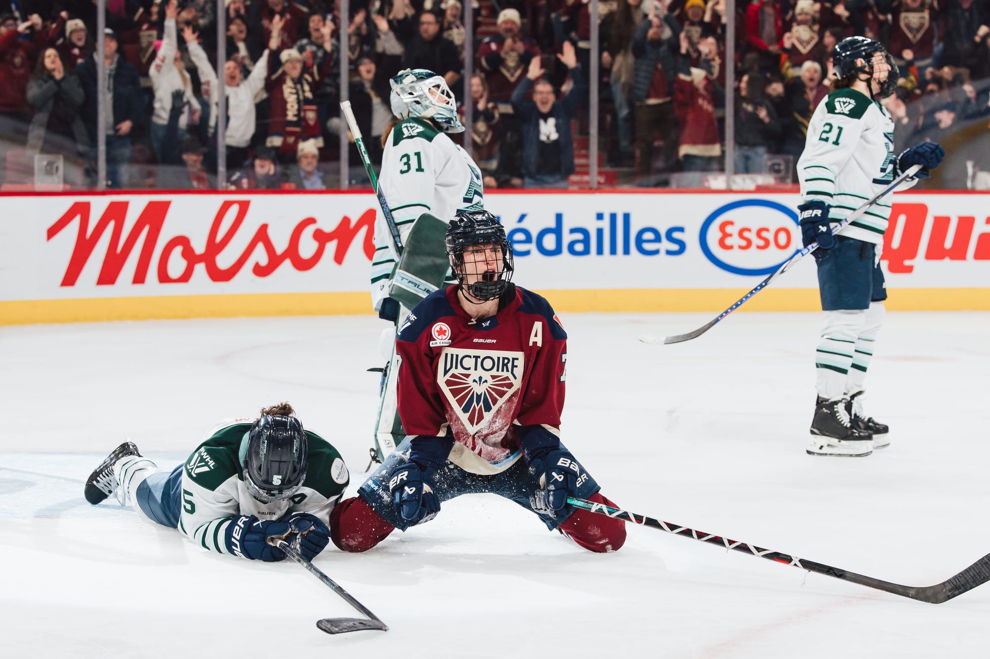 Stacey kneels on the ice, yelling in celebration. She is wearing a maroon home uniform. Frankel and two Fleet skaters are around her looking disappointed. 