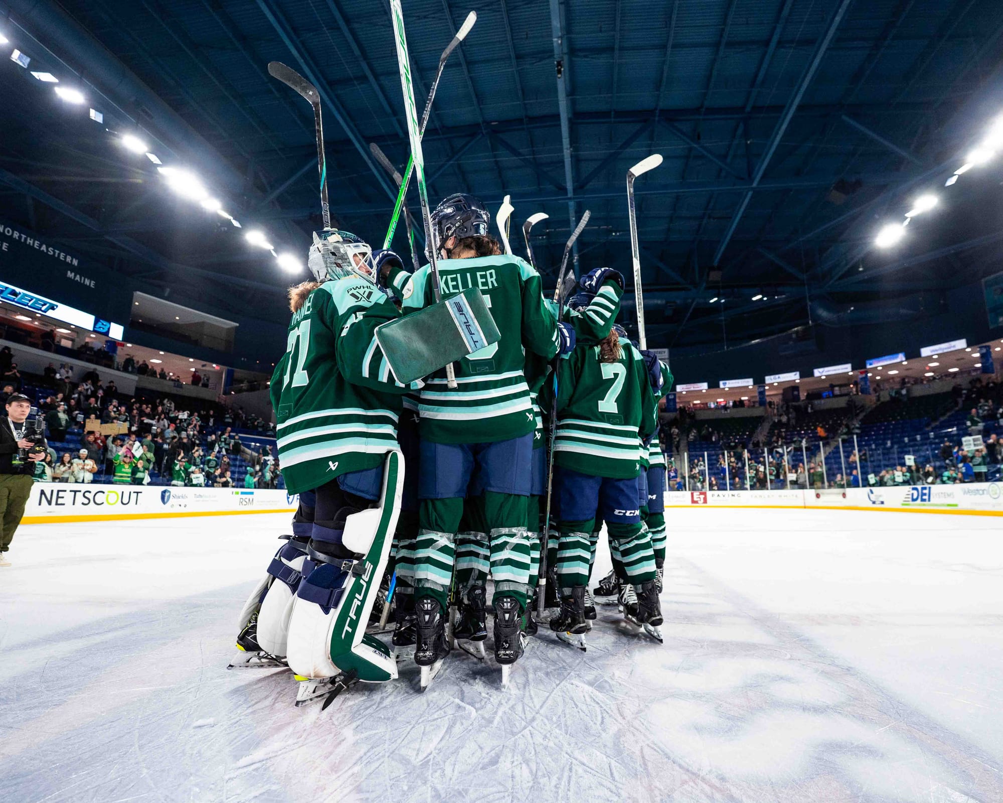 Fleet players raise their sticks and celebrate with a group hug at center ice. They are wearing green home uniforms.