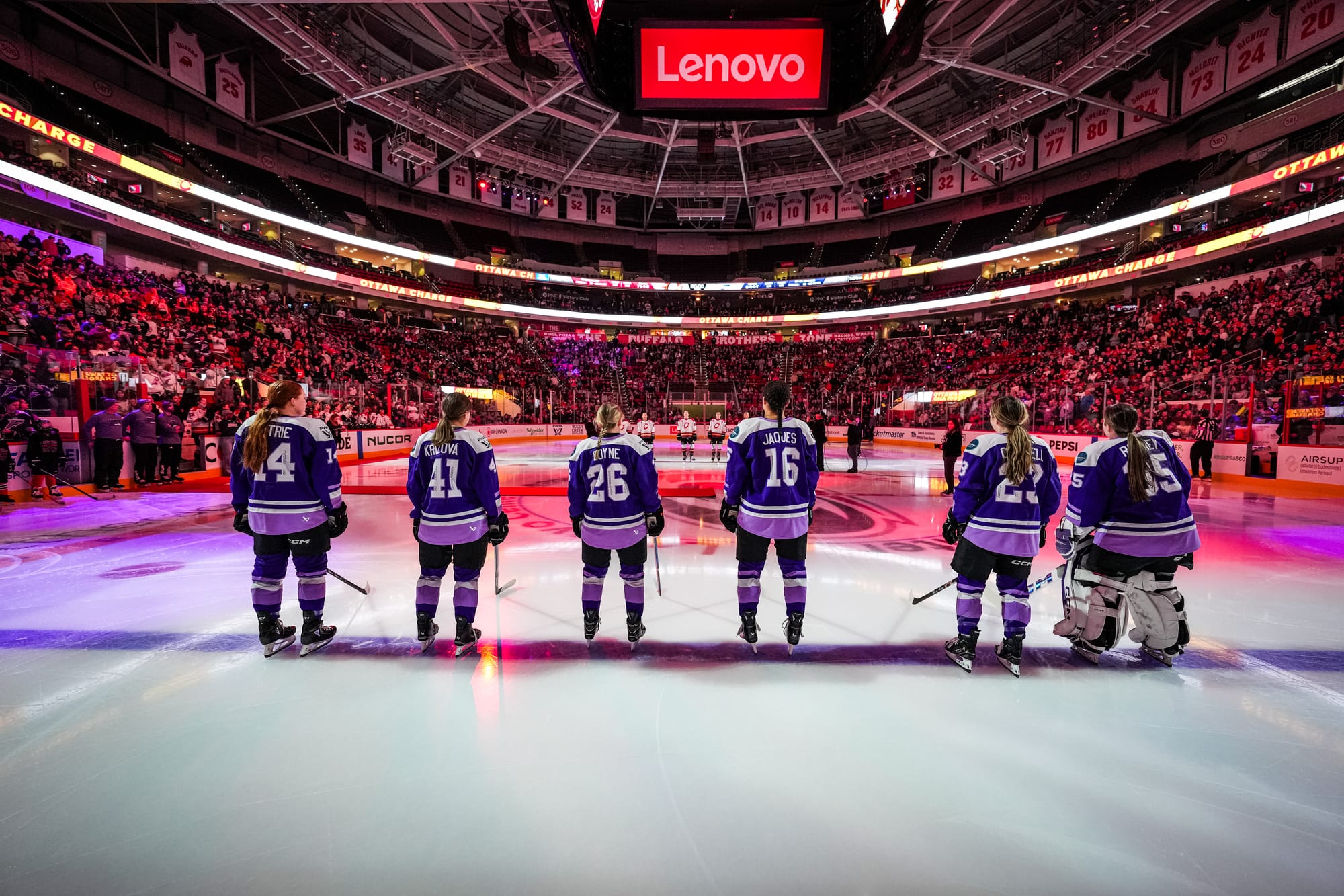 Frost skaters line up along the blue line during pregame proceedings. Their backs are to the camera, and they are wearing purple home uniforms.