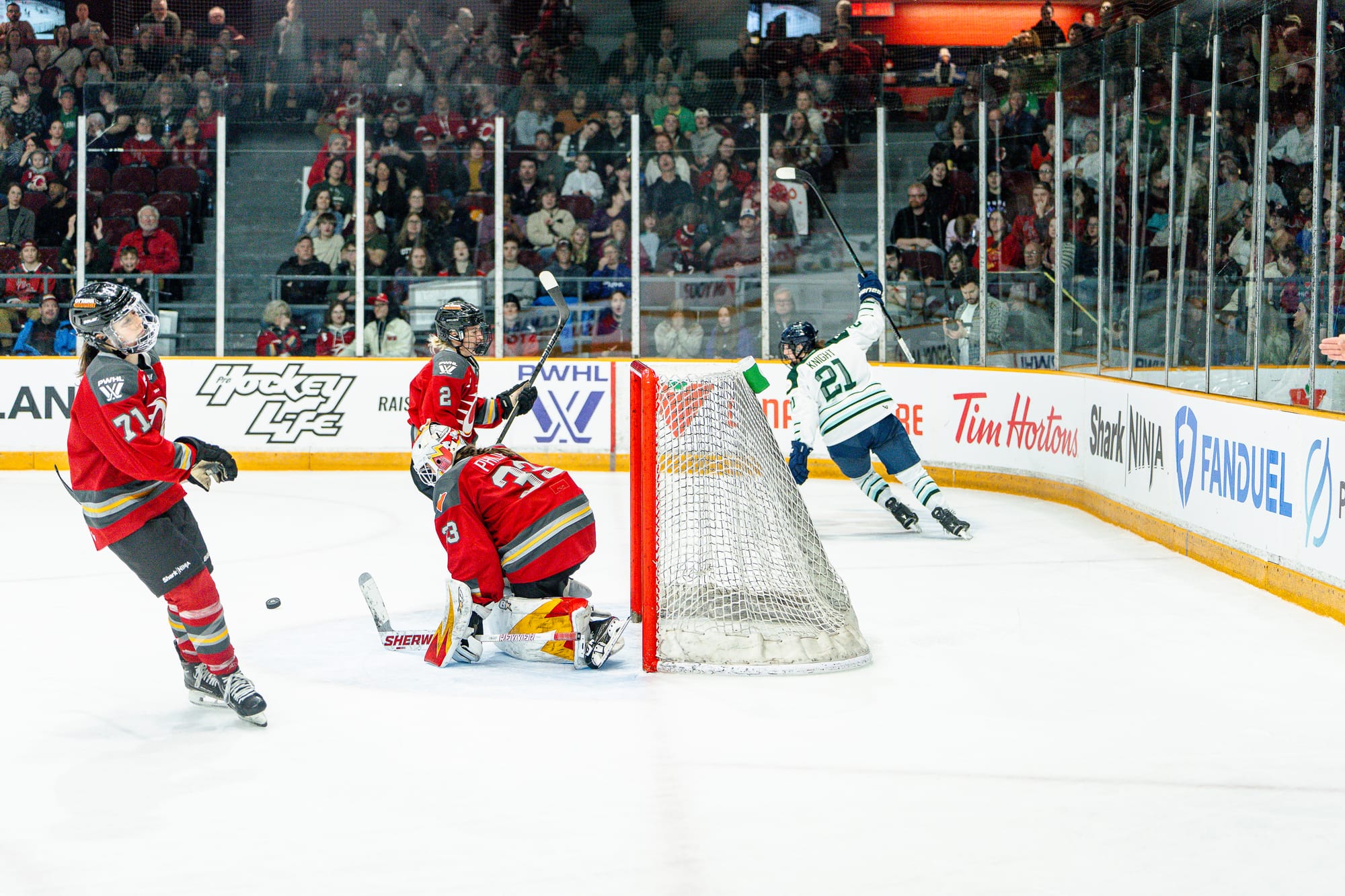 Knight raises her stick as she skates away from the goal. Phillips is on her knees in the crease, while two Charge players skate away disappointed. Knight is in a white away uniform, while the Charge players are all in red home ones.