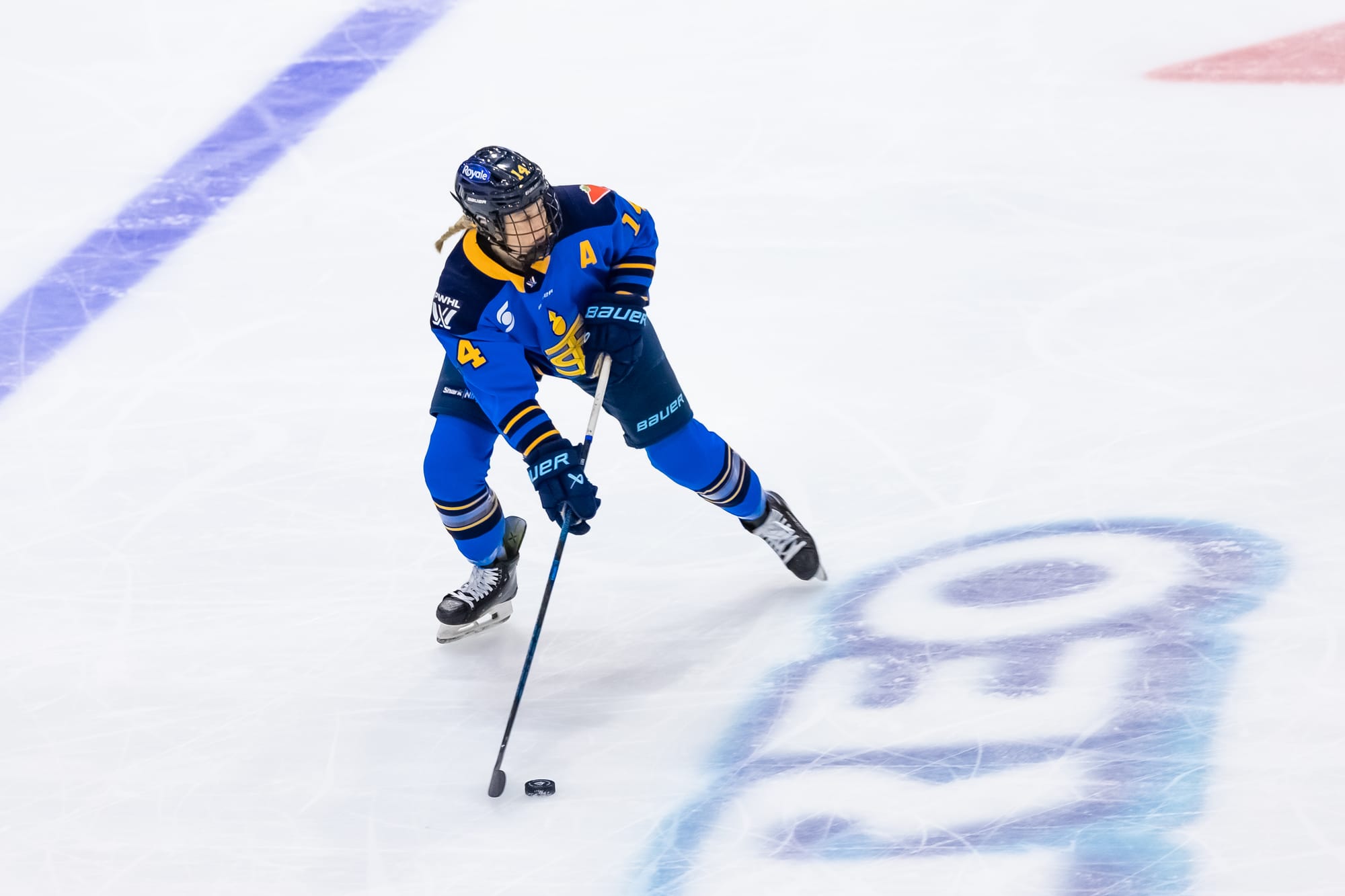 Fast looks up ice as she skates with the puck out to her right. She is wearing a blue home uniform.