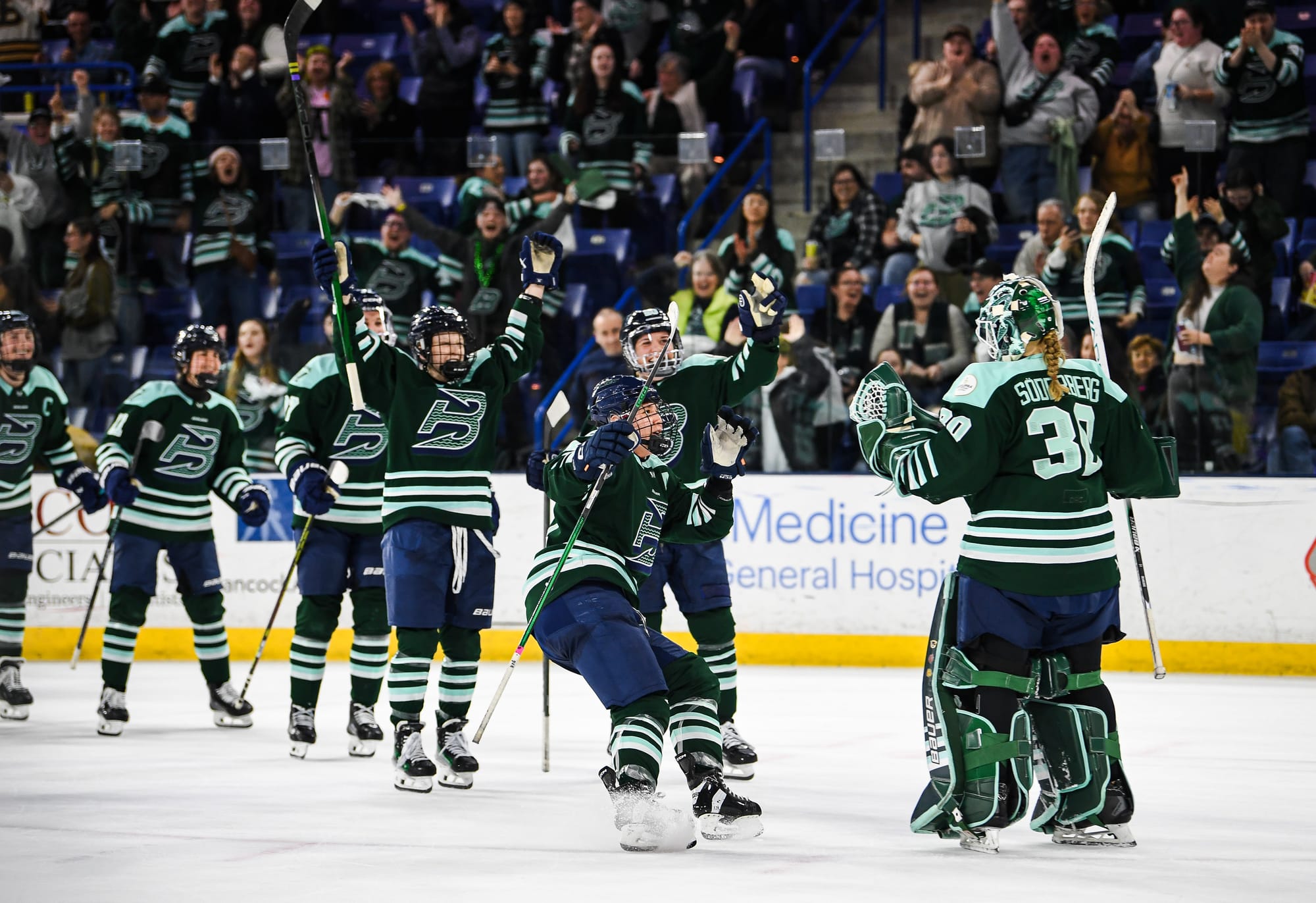 Fleet players smile and skate to hug Emma Söderberg, who is pumping her fists in celebration. They are all wearing green home uniforms.