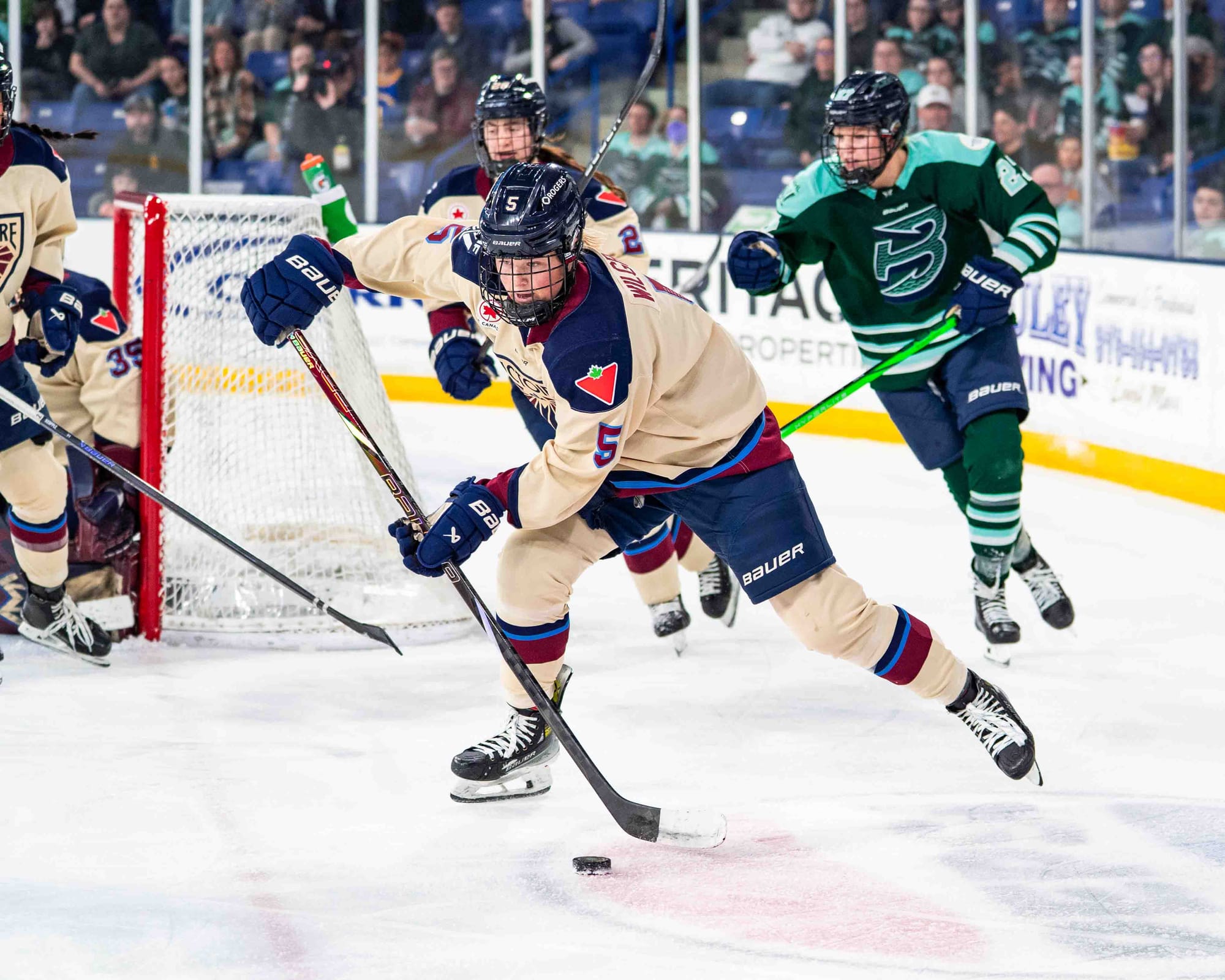 Wilgren skates the puck out from behind the net, and she appears to be preparing the pass it. She is wearing a cream away uniform.