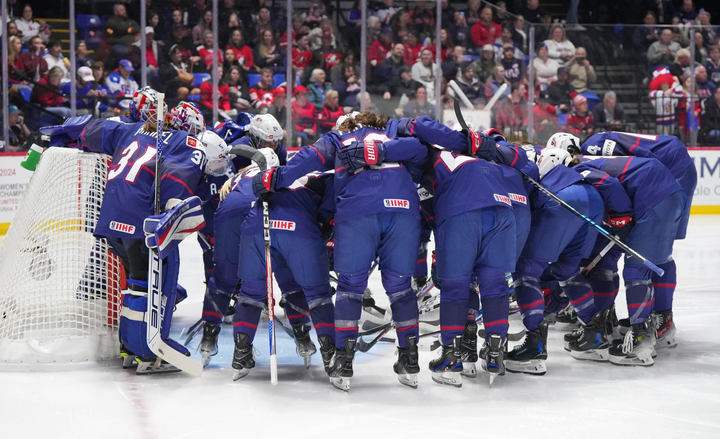 All members of Team USA are arm-in-arm, hunched over in a circle in front of their goal. They are wearing blue uniforms.