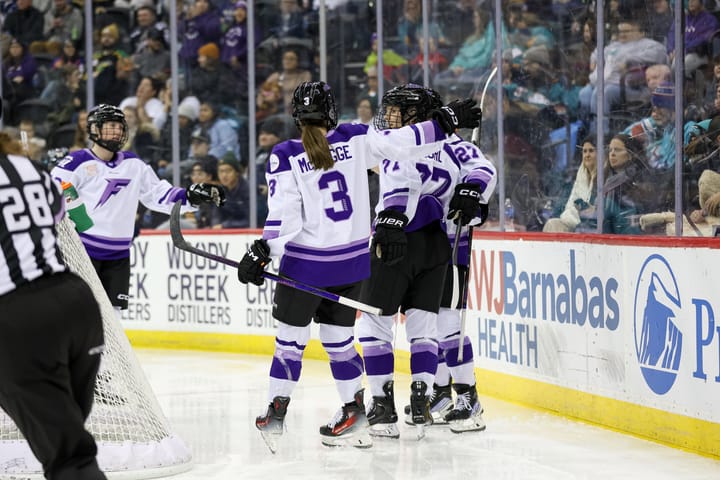 Four Frost players celebrate with a group hug behind the net. They are wearing white away uniforms.