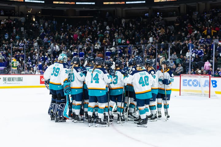 A from-behind look at the entire Sirens team in a tight huddle. They are wearing white away jerseys.