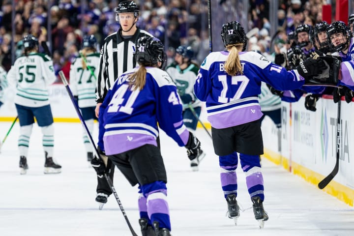 Bryant and Křížová skate down the handshake line at the bench, backs to the camera. They are wearing purple home uniforms.