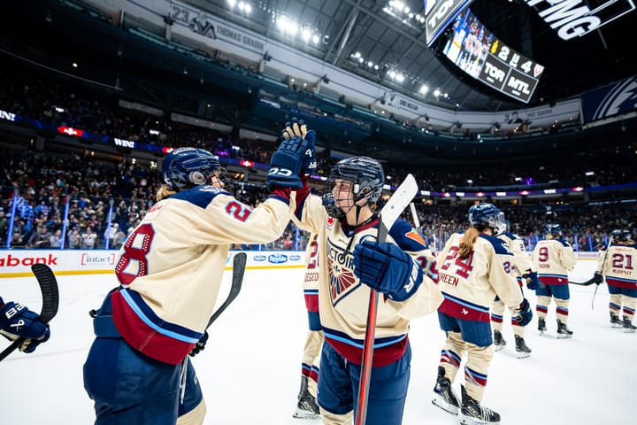 Poulin and Dubois high-five. Other players skate away having just high-fived Poulin. They are wearing cream uniforms.