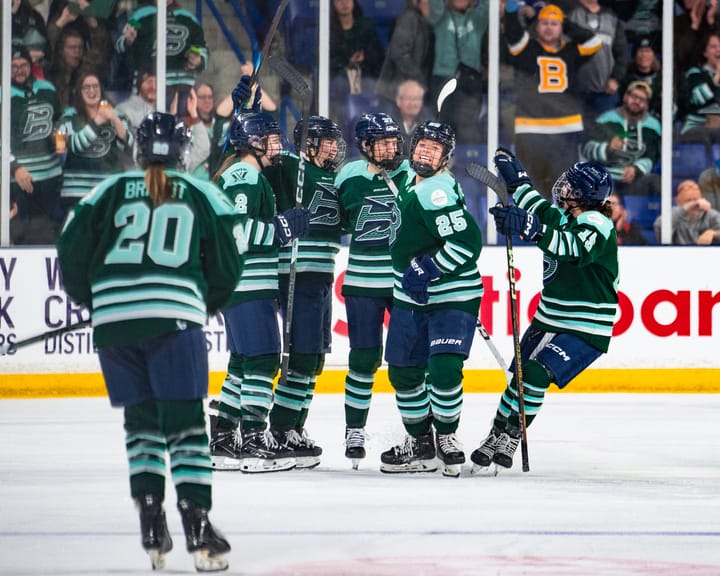 Five Boston Fleet players celebrate with a group hug. Hannah Brandt is skating to join. They are wearing green home uniforms.