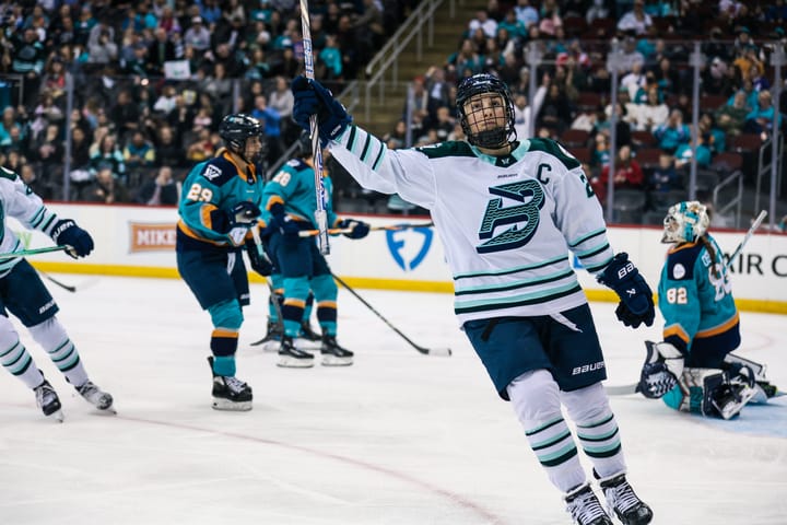 Knight raises her stick in celebration. She is facing the camera and wearing a white away uniform.