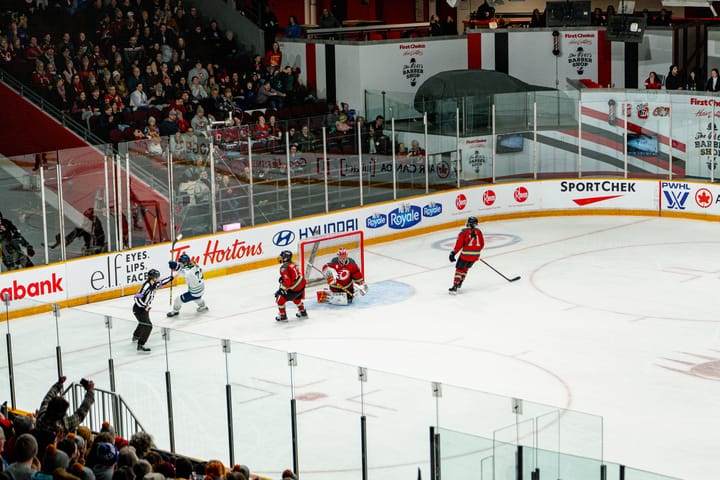 An above-ice view of Tapani raising her stick in celebration as the ref points to the goal, as two Charge players skate away.