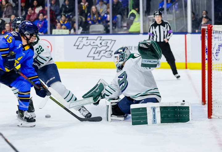 From her knees, Frankel pushes to the side and kicks the puck out to her right. She is wearing a white away uniform.