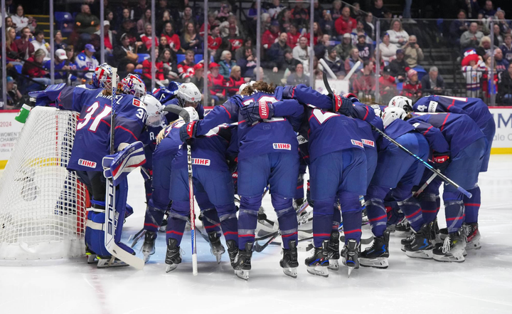 Team USA locks arms and leans over in a huddle in front of the goal. They are wearing blue USA uniforms.